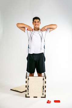 A boy demonstrates the use of a piece of exercise equipment, the 'Gym Box'.  He is standing within the box, pulling upwards on resistance bands that are attached to either side of the box.