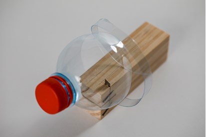 A wooden cutting tool that cuts plastic bottles. This is a small wooden block with a sharp blade embedded within it, being used to cut a spiral shape out of a plastic water bottle with a red cap is shown being spiral cut