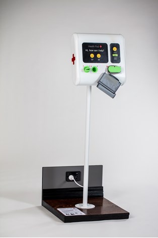 An overall view of a white structure that holds a screen mounted on the pole. The screen has a red medical cross on the side,  with green buttons and a yellow sad and happy face. Instructions are provided on the base of the object.