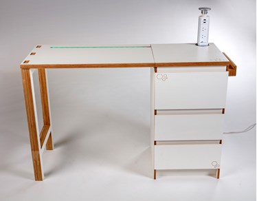 A front on view of a white and wooden desk. The desk is supported with two legs on the left. Three drawers support the construction on the left. A powerboard can be accessed on the right. 