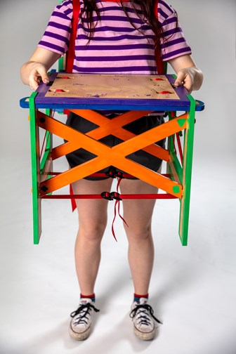 A girl in colourful clothing is holding a multi-coloured wooden stool.  This has a purple top, orange sides and green legs.