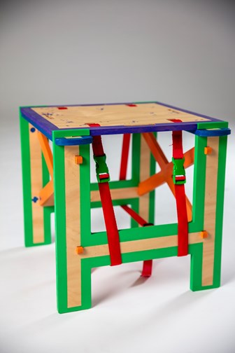 A square multi-coloured wooden stool. The legs are green, the top is purple and the side bracing is orange. It has red nylon straps on either side.