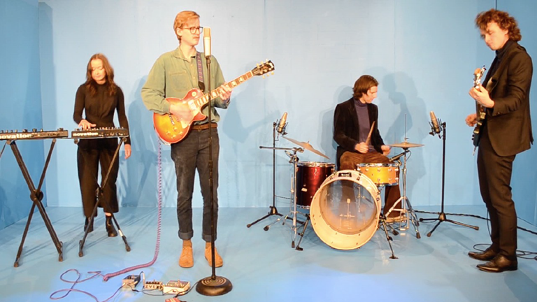 One woman playing synth, and three men playing the guitar, drums and bass in a blue room