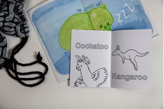 An opened colouring book shows a black outline illustration of a cockatoo and a kangaroo