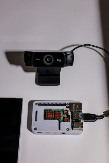Black Logitech HD 1080p webcam positioned above a Raspberry Pi board in a white case with exposed copper heatsinks and connected cables.