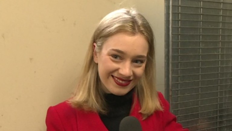 A woman with blonde hair and a red jacket stands in a corner holding a Channel 9 News microphone