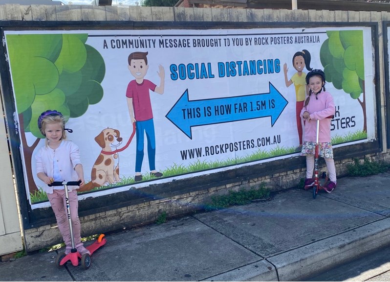 Two girls on scooters standing in front of large 'social distancing' poster on a fence
