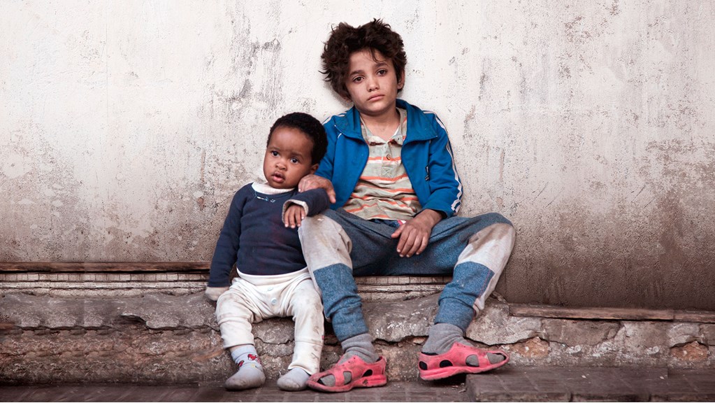 A child and toddler lean against each other on the street, looking despondent