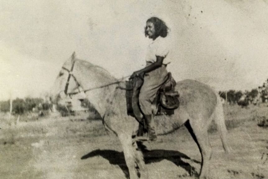A black and white photograph of a smiling young woman atop a horse. 
