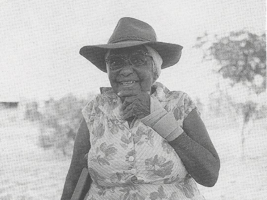 A black and white photograph of an elderly woman smiling to camera in the bush. 