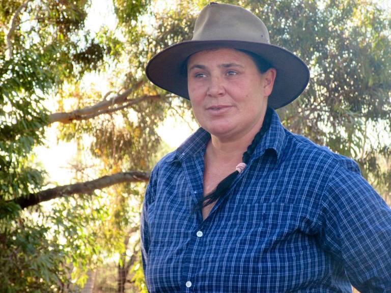 A colour photograph of a woman in an wide-brimmed felt hat and blue chequered shirt amidst trees.