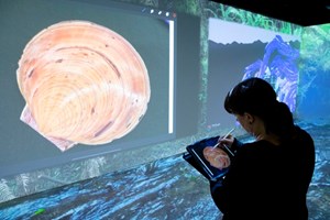 A woman drawing a shell on an iPad that is projected in large-scale onto the walls of a digital immersive space