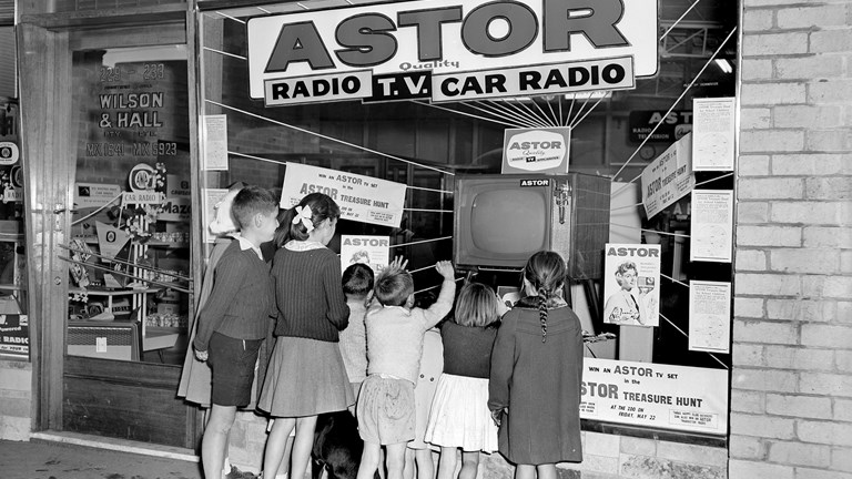 Promotional image of a group of people looking at the Wilson Electronic Store display window. There is a display of Astor radios and televisions.