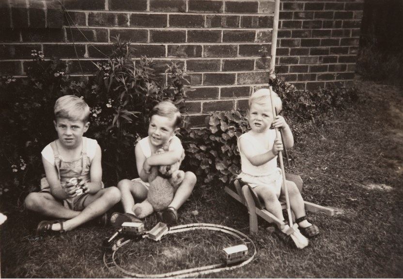 Three boys playing with a toy train set, teddy bear and other toys on the lawn of a backyard.Three brothers Darryl, John and Trevor Hawksworth, in the backyard of the Hawksworth family home, Strathmore, 1949.