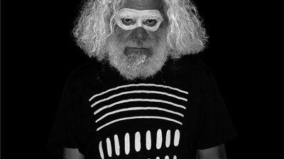 A man with white hair, beard and paint around his eyes stands wearing a black shirt that blends into the black background. White horizontal and vertical stripes are visible on his shirt.
