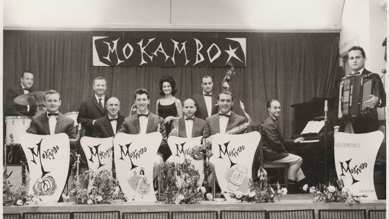 Eleven Mokambo Orchestra band members, including a female singer. Band leader Ugo Ceresoli with accordion at far right. There are six music stands standing in front of band members across the front of stage.
