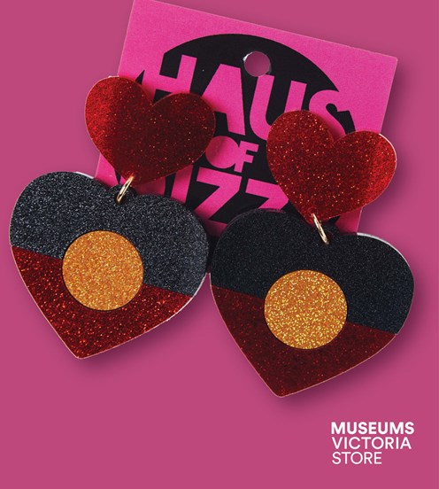 Flay lay image of a pair of heart shape earrings depict the Aboriginal flag on a pink background
