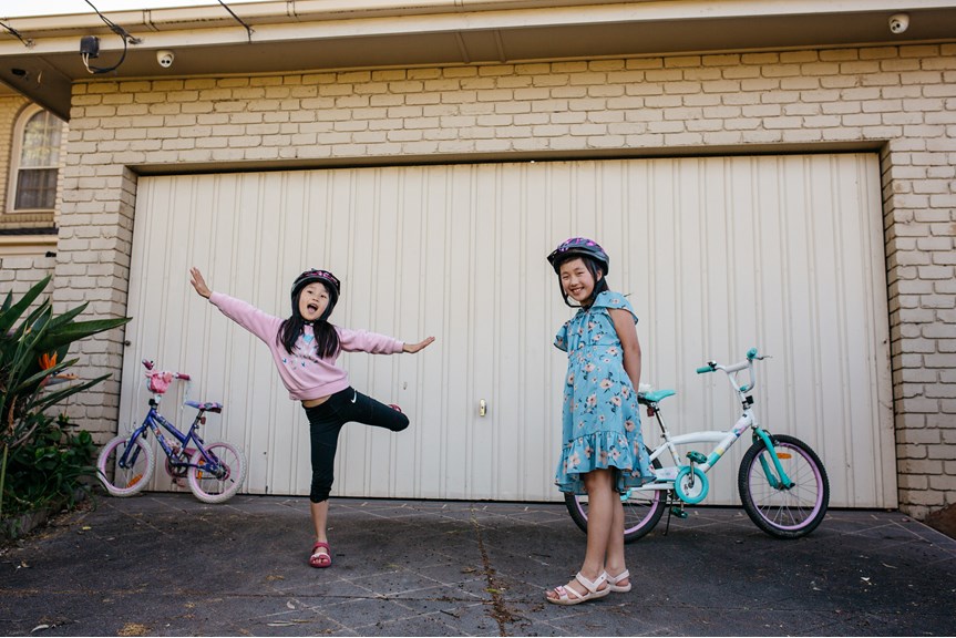 Two girls wearing bike helmets standing in front of their family home. Their bikes are leaning on the garage door
