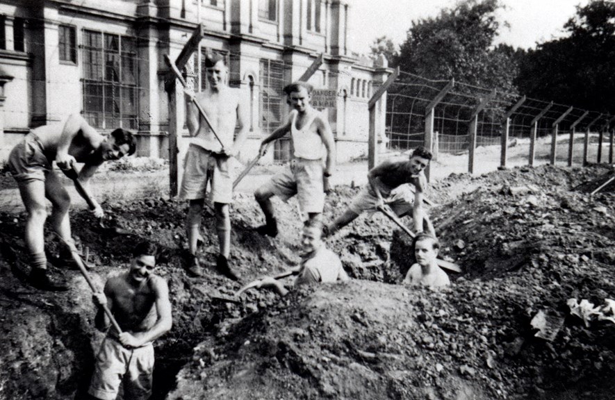 Black and white photograph of seven young men, smiling, wearing military shorts, some in white singlets, and some with bare chests digging a trench.