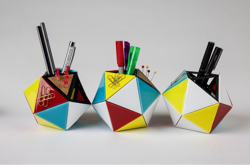 Three modular stationery holders are lined up on a white background. The holders are comprised of red, white, blue and yellow triangles. There are also magnetic and cork triangles holding paperclips and pins.