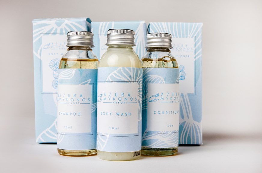 Three travel-sized shampoo, conditioner and body wash bottles are grouped in front of their individual packaging boxes. The labels are light blue with coral-like patterns. 
