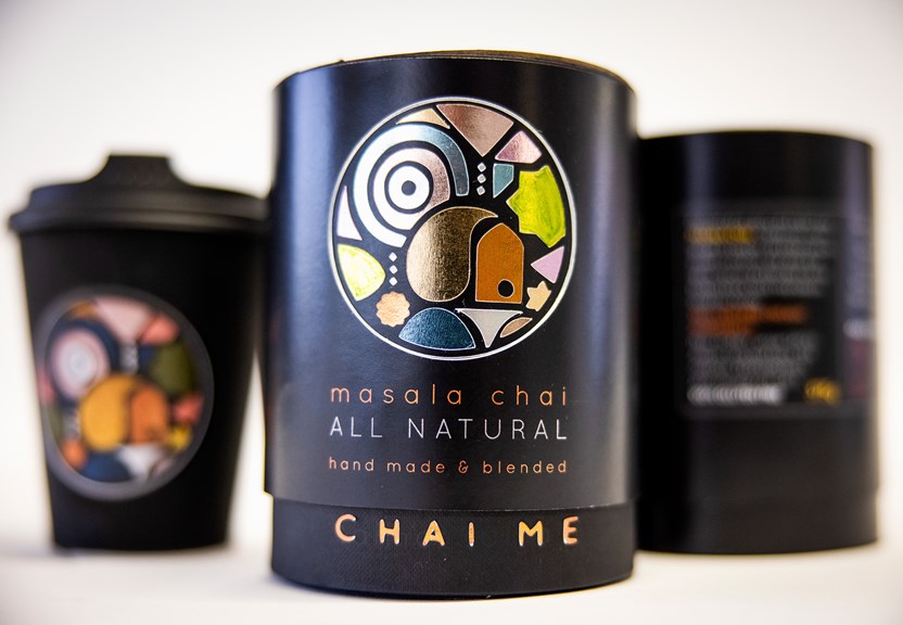 A disposable coffee cup and two cylindrical packages designed to hold loose chai leaves are featured. The packaging has the Chai Me logo which is made up of dusty colours and irregular shapes.