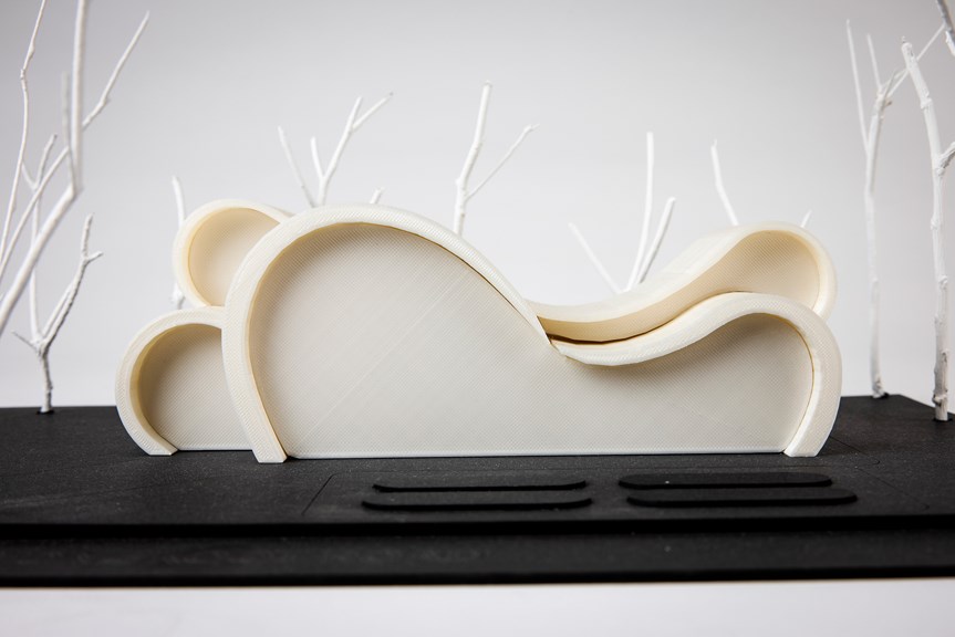 A close-up of a white architectural model, surrounded by white stick-like trees, sits on a black base. The outline of the model is defined by curved and rounded lines.