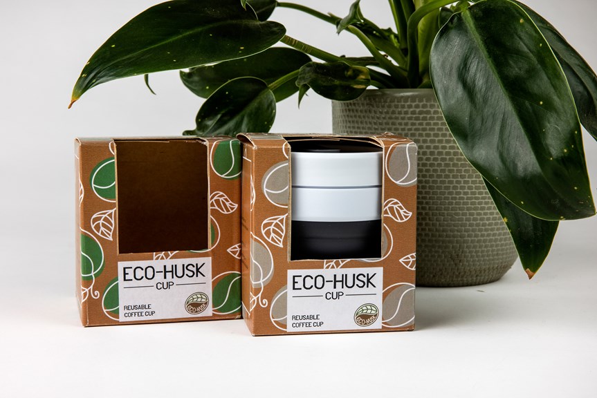 Two brown boxes sit in front of a plant in a green ceramic planter. The boxes are branded Eco-Husk with green and grey leaves. A reusable coffee cup sits in one of the boxes.