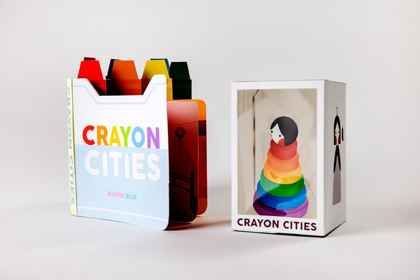 A board book is displayed fanned open. Each page has a tab designed to look like a different coloured crayon. To the right sits packaging for a stackable toy. 