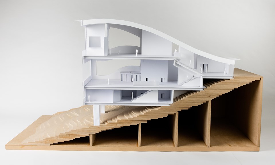 A photograph of an architectural model with three levels and built into a hill. The roof structure is curved like a wave. The side wall is removed, showing the layout of each of the floors.