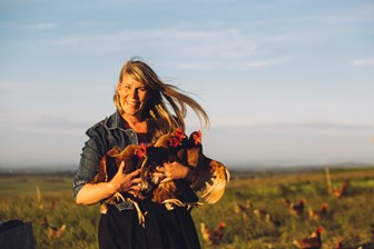 Woman standing in a field holding four chickens in her arms, other chickens can be seen grazing on the grass