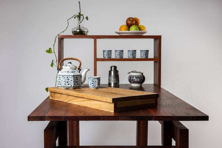 The image shows the Tea Table from one side. It is made of dark wood. Its inbuilt shelves are visible, and are holding tea cups, fruit, and a potted plant. There is a traditional style teapot on the table.