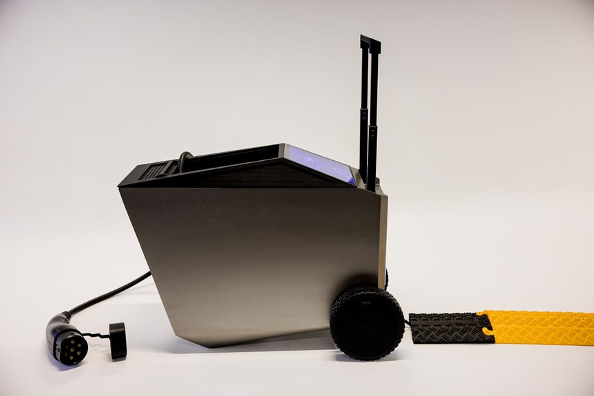 The image shows the portable electric vehicle charger from a side view. It is a modern, geometric shape with a metallic finish. Its wheels and handle are visible, and there is a charging cable coming out of the top.