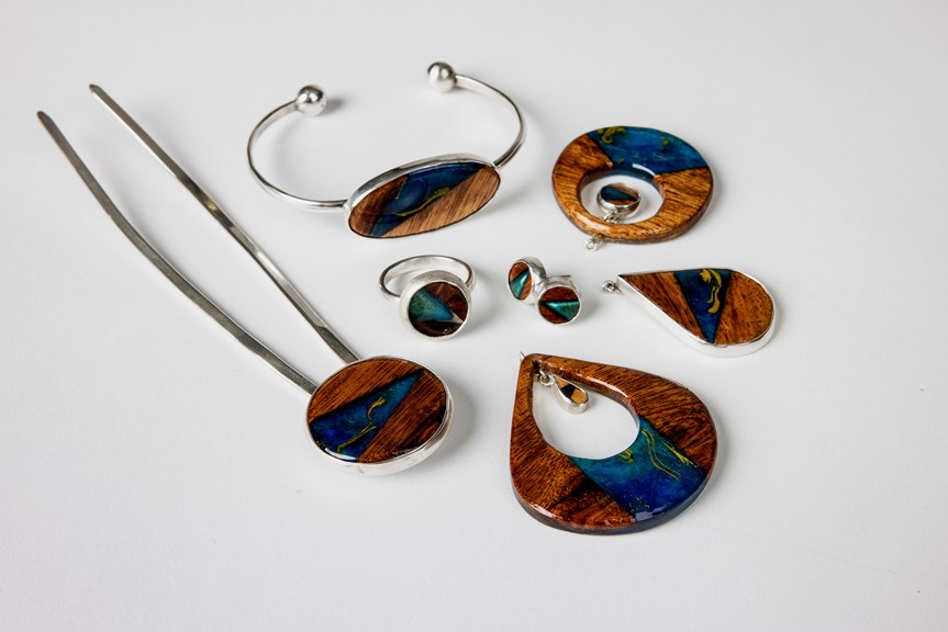 Six items of jewellery are shown lying on a white surface. The pieces incorporate circular forms with wooden and blue resin materials and are set in silver.