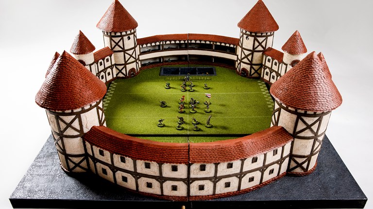 The ‘Blood Bowl’ Stadium is shown in full, with several miniature figures arranged on the stadium’s fake turf, as if a game was underway. The stadium is circular, with Tudor style walls and turrets.