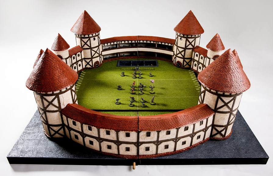 The ‘Blood Bowl’ Stadium is shown in full, with several miniature figures arranged on the stadium’s fake turf, as if a game was underway. The stadium is circular, with Tudor style walls and turrets.