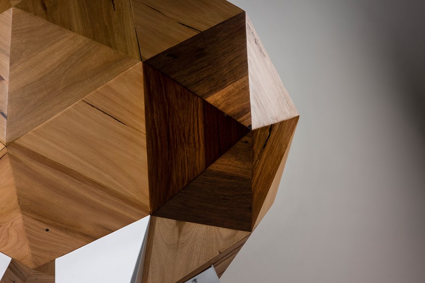 A close-up of the chair, depicting its triangular geometric shapes. It is made of wood, and the light is reflecting on its panels.
