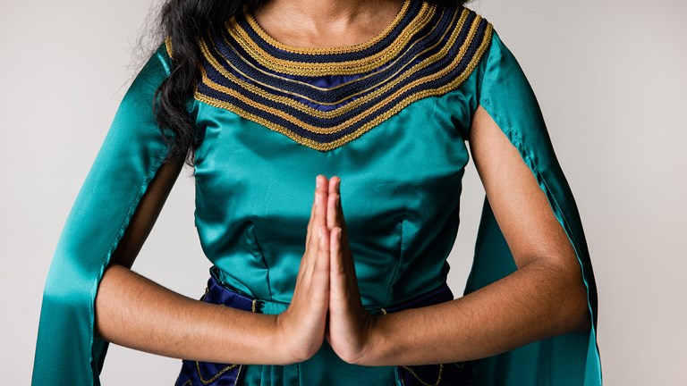 A person with long, dark hair is shown wearing a garment made from emerald green silk. The garment has gold and navy blue embellishments around the neckline. The wearer is holding their hands together as if in prayer.