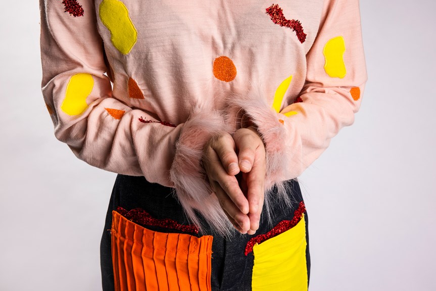 A person wears a light pink, long sleeved top with faux fur trim on the sleeves. Various geometric shapes adorn the top, made from different fabrics. The person also wears a pair of denim pants with colourful patches on both legs.