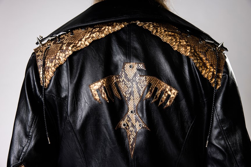 The back of a black leather coat is pictured, which has an eagle emblem in the center made out of snakeskin. There is also snakeskin on the shoulders of the coat, with some spike embellishments and lacing.
