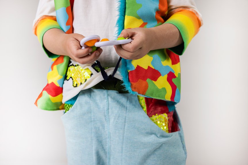 A person wears a multicoloured jacket with a blue feather trim. They hold plastic toys with tactile features that are attached to the drawstring of a pair of pants. The pants have colourful pockets adorned with sequins and furry fabrics.