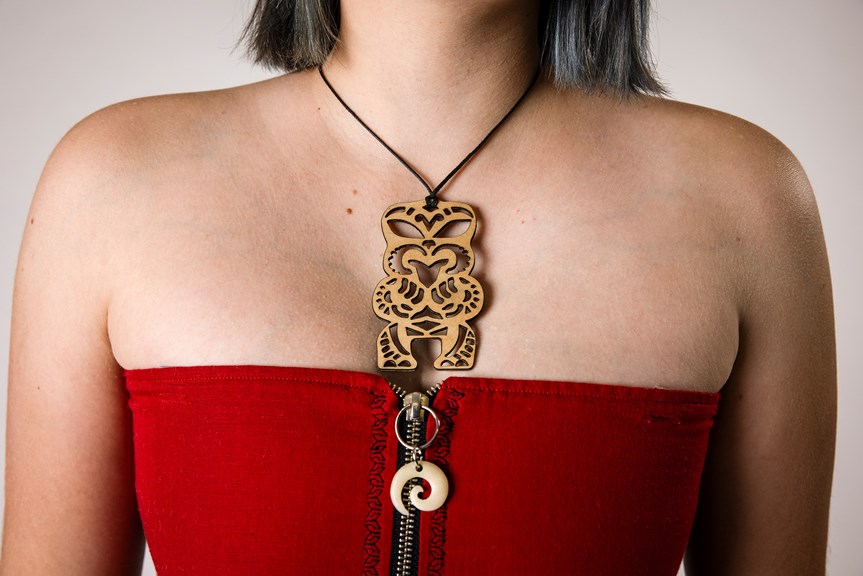 A person is shown wearing a necklace that has a large Tiki pendant made of plywood. They also wear a sleeveless red top with a zip in the centre, which has a circular bone adornment on its closure.