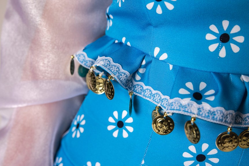 A close-up of several small gold coins, shown hanging from a laced hem on a garment. The garment is made of a blue fabric with a modern floral pattern.
