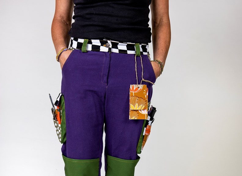 A person is seen front-on, wearing a pair of purple and green trousers with a checked waistband. The trousers have large exterior pockets on each side which are holding various small tools.