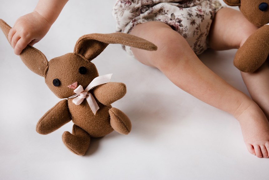 A baby’s hand is in the picture, holding onto the ear of a toy rabbit. The rabbit is made of brown felt, with buttons for eyes and a silky pink ribbon around its neck.
