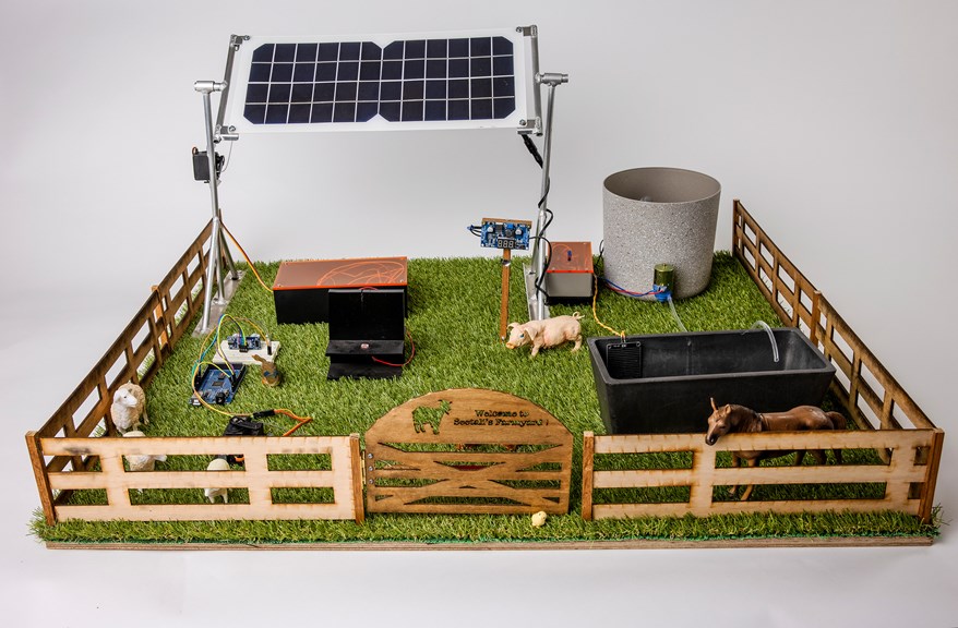 An image of a model of a farm. It is surrounded by a fence which contains a solar panel, a water tank and a water trough, and toy farm animals.