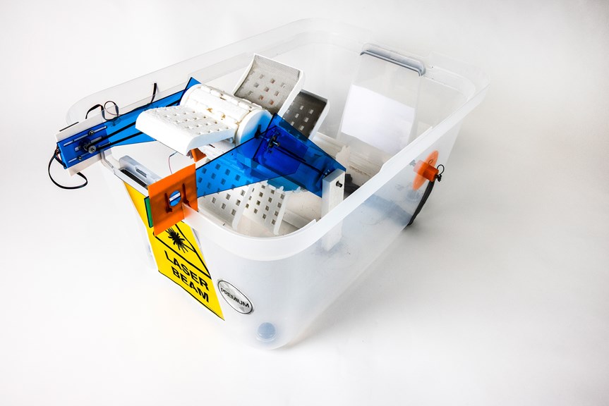 A clear plastic tub has a yellow label reading “laser beam”. Inside the tub are various motorized components, most notably a conveyer belt system. Blue and orange acrylic arms secure the belt.