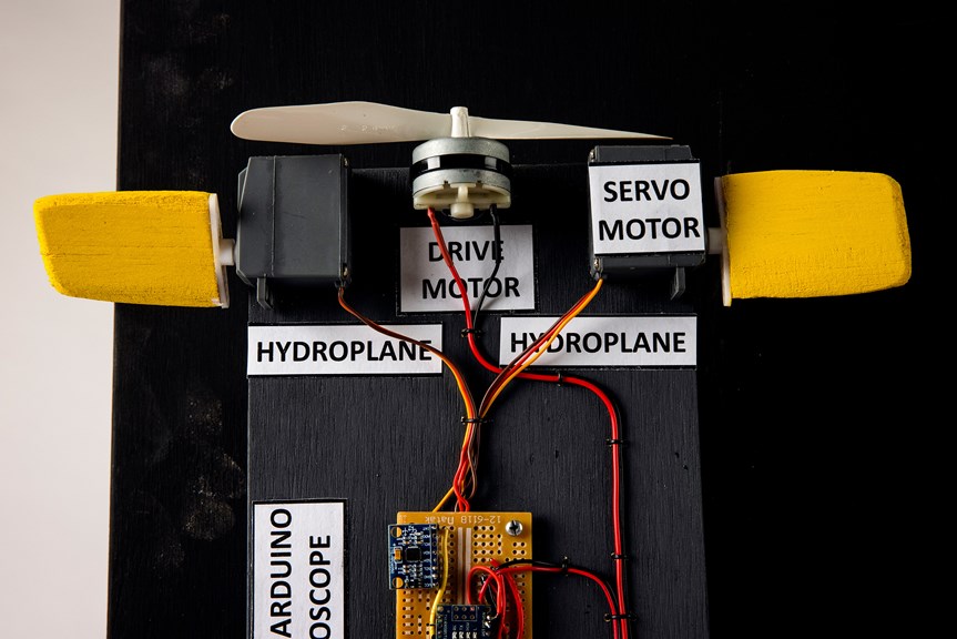 A close-up image of the Autonomous Underwater Vehicle is in shot, depicting its motorized components. There are red and yellow wires running from an Arduino to a propellor. The components have text labels, reading “hydroplane”, “drive motor”, and “servo motor”.