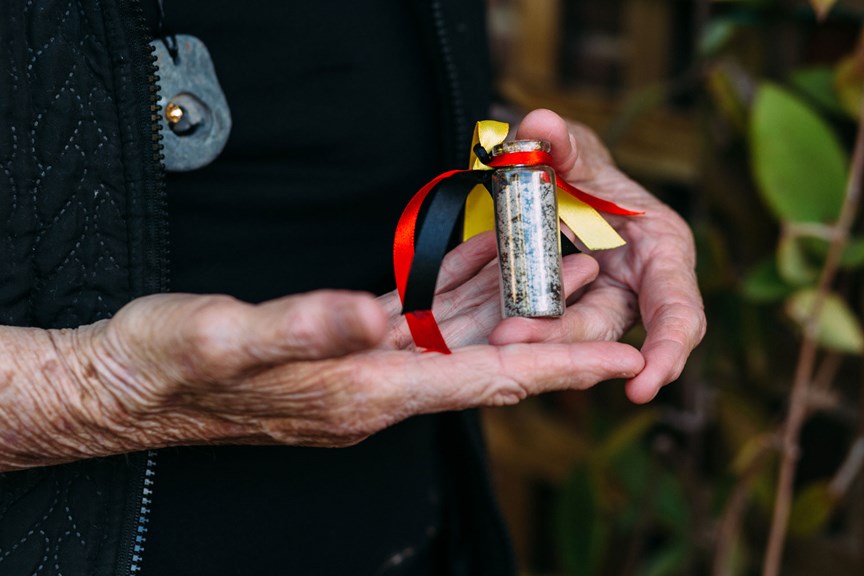 Hands holding a small viral of soil with the ribbons in the Aboriginal flag colours tied to the top