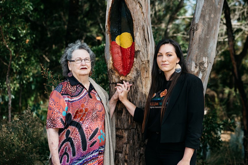 Two women holding hand standing in front of a scar tree with the aboriginal flag paint on the scar